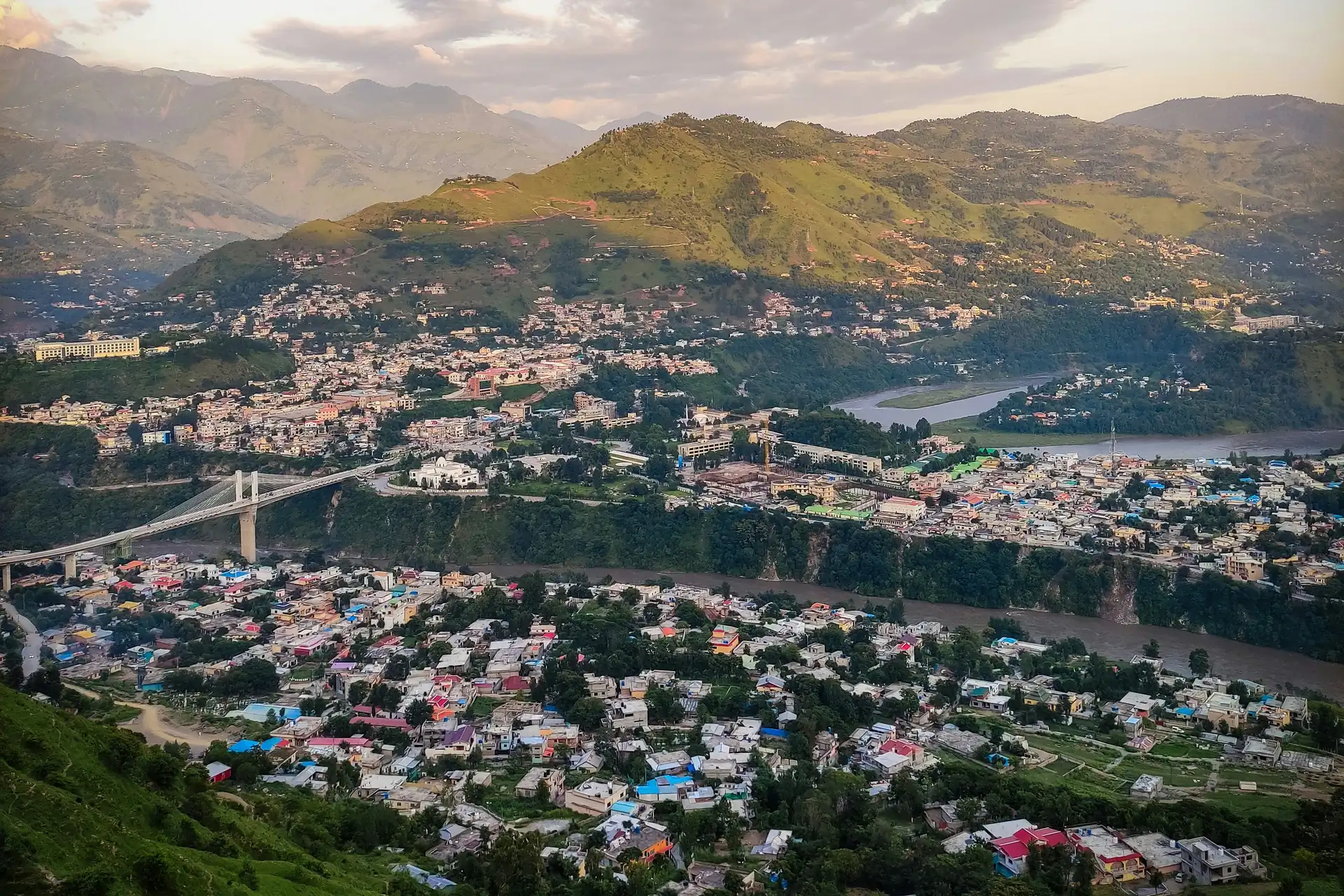Muzaffarabad in Azad Kashmir seen from a hillside, the river and a bridge running through the city below green hills.
