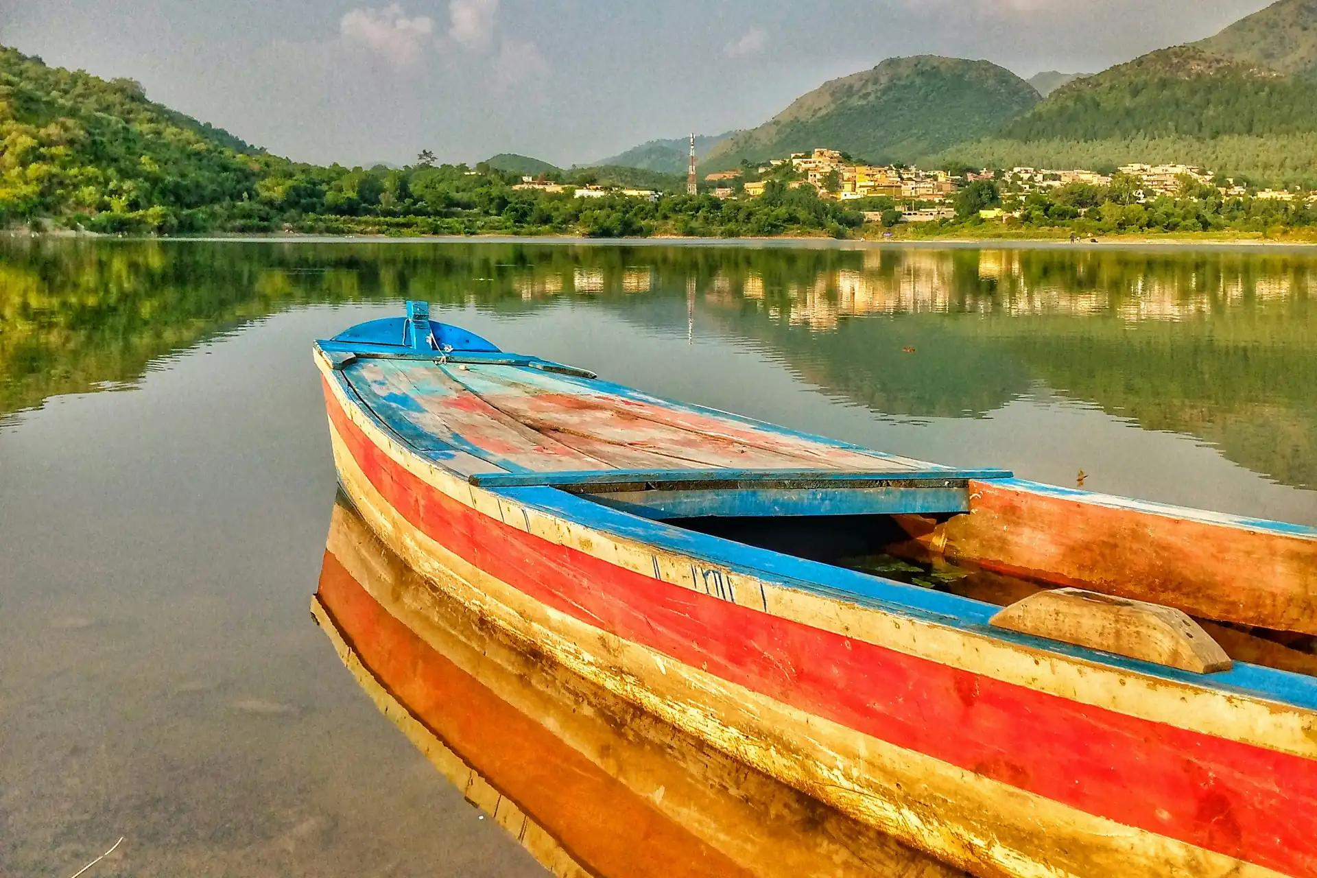 A painted wooden boat on the still water of a dam in Haripur, with green hills behind.